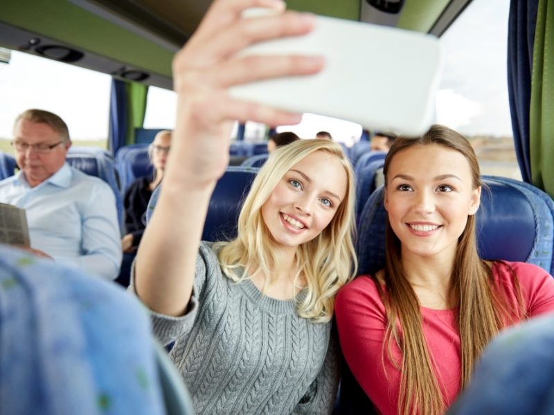 transport, tourism, road trip and people concept - happy young women or friends in travel bus taking selfie by smartphone